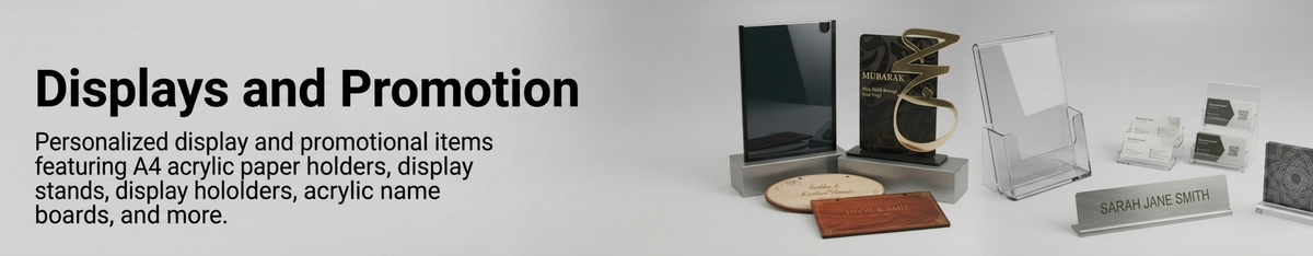 Promotional display set featuring personalized acrylic paper holders, display stands, a black plaque on a gray base, a black award-style book with a gold ribbon, a round wooden plaque, and a small wooden box, arranged on a light gray background with the heading 'Displays and Promotion'.