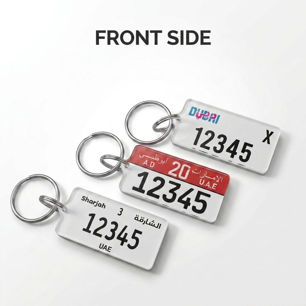 Three rectangular license plate keychains shown from the front on a light gray background, each attached to a silver metal key ring. The top keychain is white with blue and pink 'Dubai' text and black numbers; the middle keychain has a red header with white text and black numbers; the bottom keychain is white with black text.