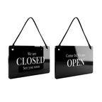 Two glossy black hanging signs with black cords, shown side by side on a light gray studio background. One sign reads 'We are CLOSED See you soon' and the other reads 'Come In! We are OPEN'.