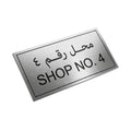 A rectangular brushed-metal sign with black Arabic text above the English text 'SHOP NO. 4', shown floating at an angle on a white background.